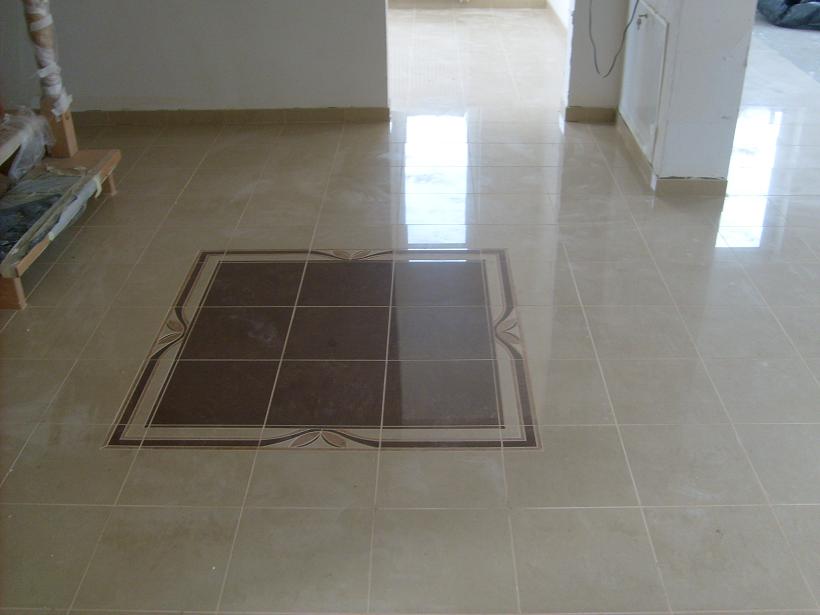 Ułożona beżowa podłoga z płytek ceramicznych z dekoracyjnym, kwadratowym wzorem z ciemnobrązowych płytek na środku pomieszczenia.